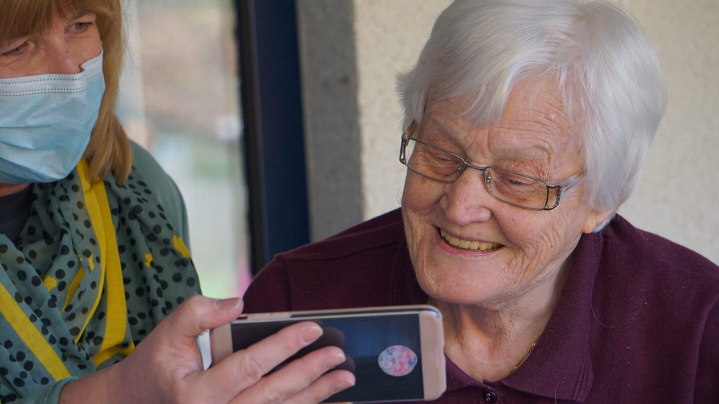 An older woman smiles as she is shown something on a phone by a younger individual, who is wearing a surgical mask.