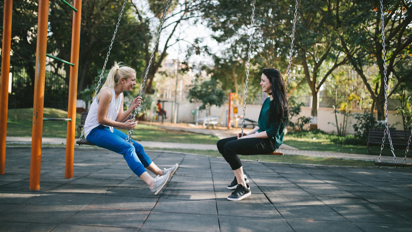 Two women in a park on swings, talking to eachother.