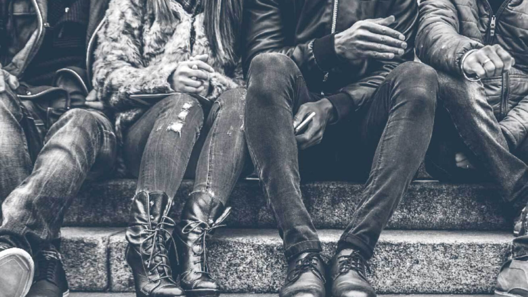 A group of young people sitting on a bench; only their legs and shoes are seen.